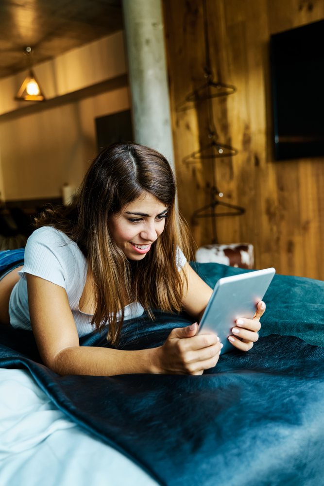 A woman smiling while using a tablet lying on a bed