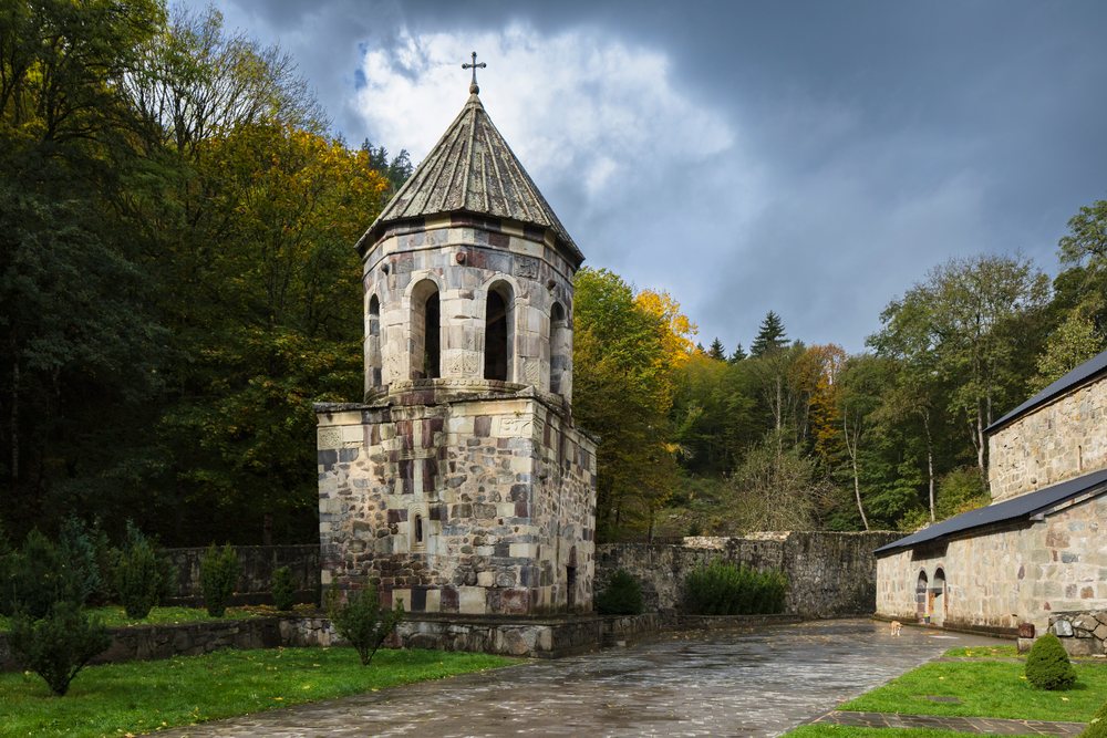 Stone bell tower with cross stands beside a stone pathway amid autumn trees at green Monastery, Chitakhevi, Georgia.