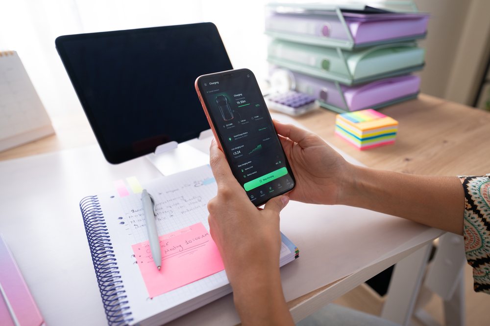 Hand holding smartphone displaying charging status beside notebook and tablet on desk in a home office, Spain.