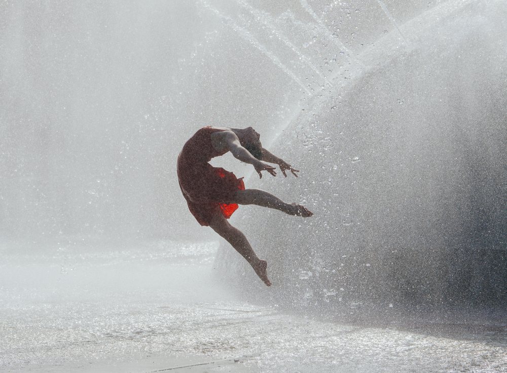 Barefoot woman in a red dress energetically jumps amidst water splashes outdoors in summer, USA.