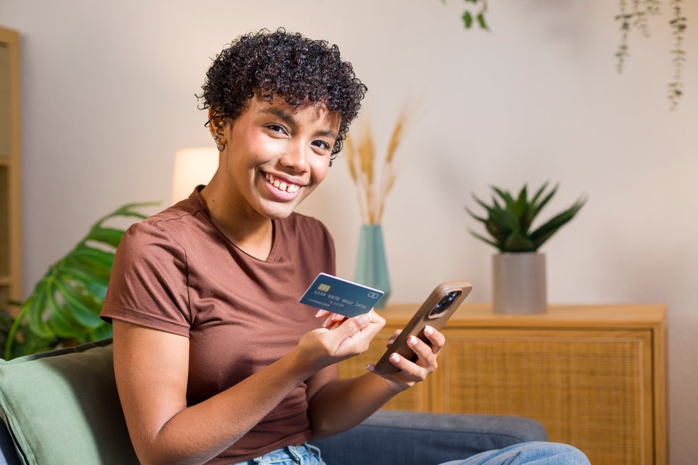 Young woman smiling and holding credit card while using smartphone, sitting on a couch in a home interior with indoor plant.