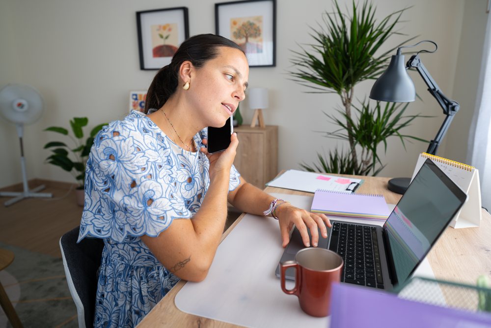 Woman telecommuting at a wooden desk in a home office, using a laptop and phone, Spain.