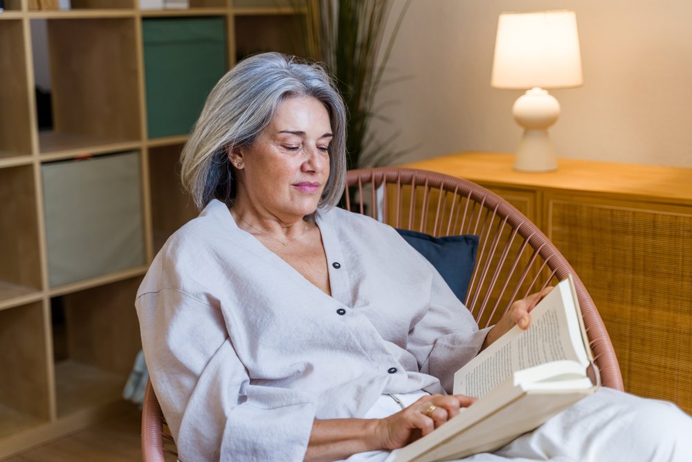 Middle-aged woman with gray hair reading a book seated in a woven round chair indoors with a lamp, shelving, and black pillow behind her, Spain.
