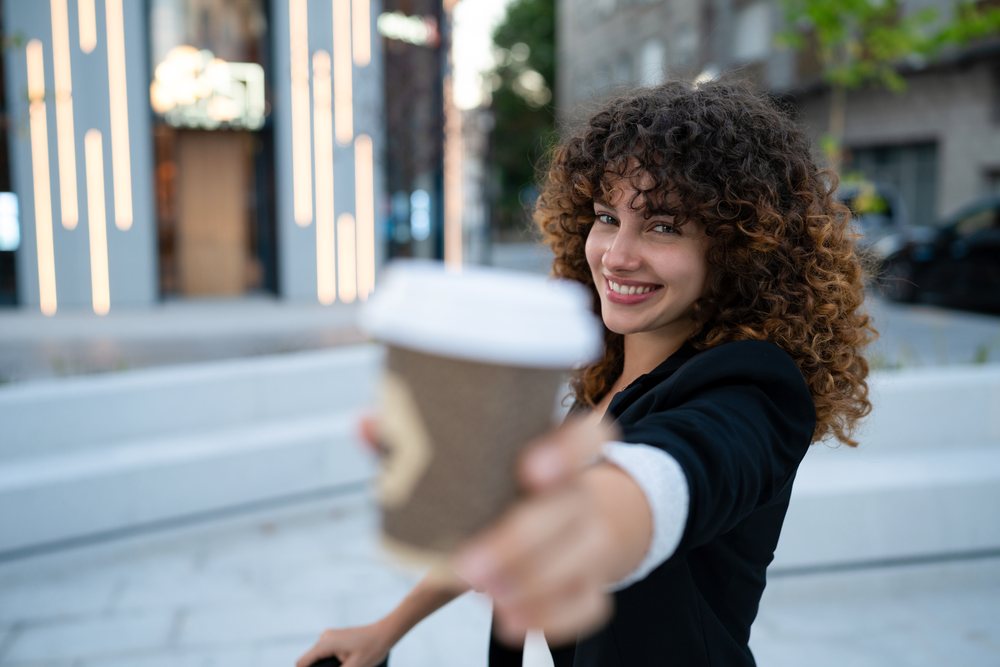 Woman with curly hair and black jacket holding a coffee cup and smiling on a sunny urban street near trees and seating, young business woman, Madrid.