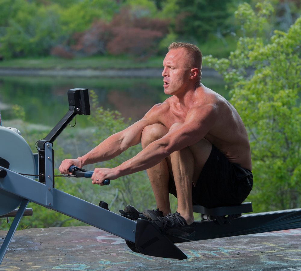 Shirtless muscular man exercises on a rowing machine outdoors near a lake, USA.