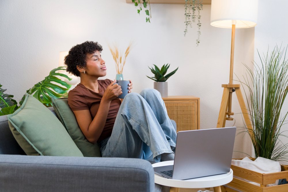 Woman sitting on a sofa drinking coffee with a laptop in a cozy living room with houseplants and modern decor, Young woman, home.
