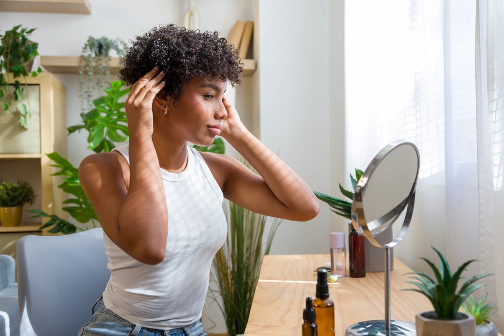 Young woman with curly hair sitting indoors at home in front of a vanity mirror with skincare bottles and potted plants in natural light.