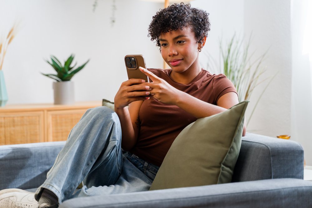 Woman with curly hair sitting on gray sofa using smartphone in living room with indoor plants and green cushion, Spain.