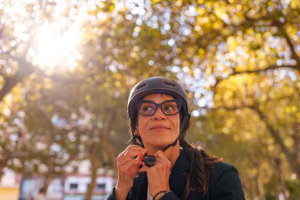Woman outdoors in a sunlit park adjusting her helmet strap among autumn leaves, Málaga.