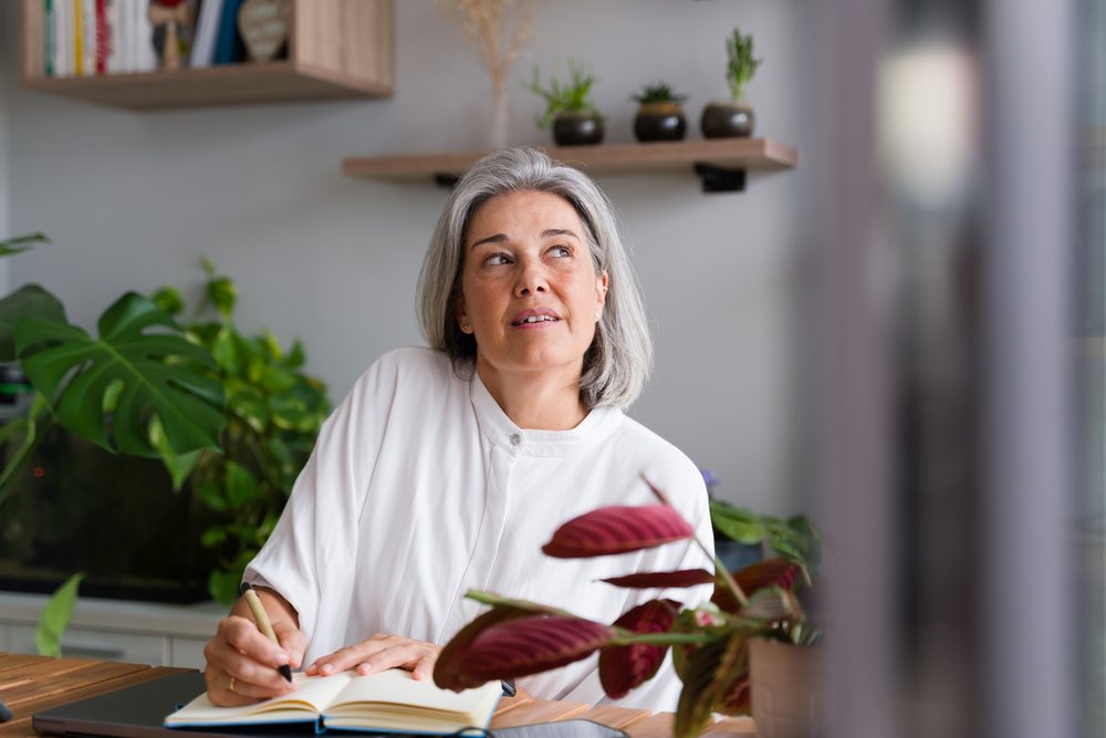Woman with gray hair writing in a notebook at a desk surrounded by indoor plants in a home office setting, Spain.