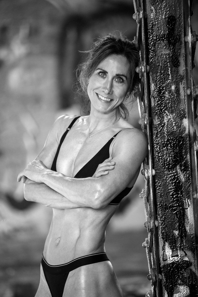 Fit woman with muscular torso and crossed arms smiling by a graffiti-covered metal beam in an urban outdoor USA setting, Calisthenics, yoga.