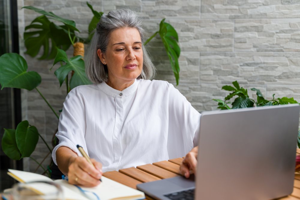 Mature woman writing in a notebook while using a laptop in a plant-filled home workspace, Spain.