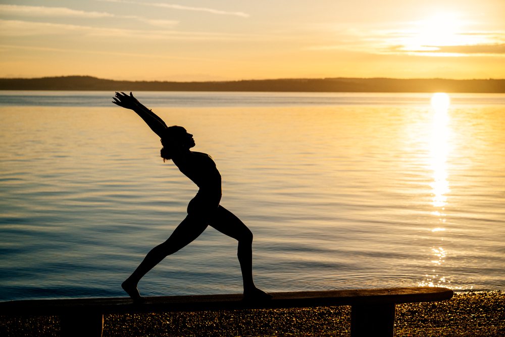 Woman performing a yoga stretch pose on a bench by a lake at sunset, USA.