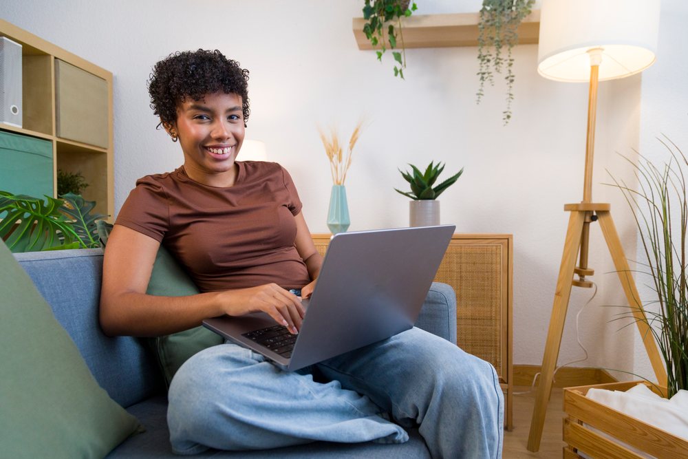 Young woman sits on a sofa using a laptop in a cozy living room with houseplants and a floor lamp nearby.