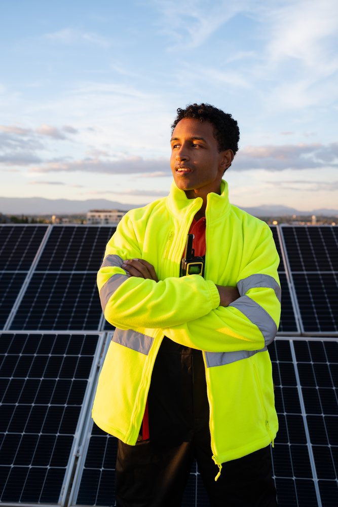 Worker with crossed arms in a reflective yellow jacket stands outdoors by solar panels at sunset, Madrid, Spain.