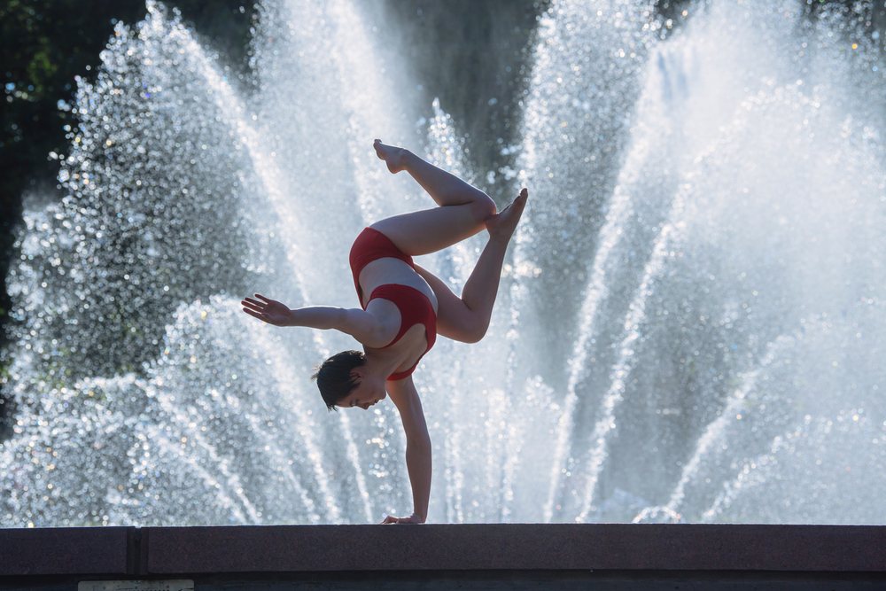 Woman in a red swimsuit performs a one-arm handstand outdoors in front of a water fountain with multiple sprays, USA.