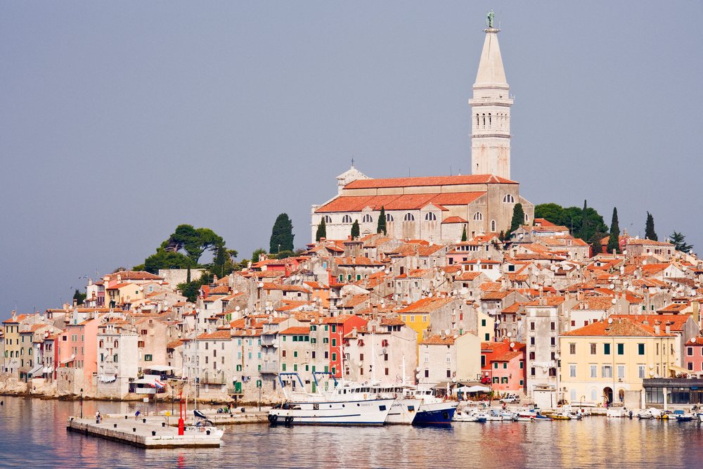 Church of St. Euphemia towers over the coastal town of Rovinj with boats docked at the pier and red tile roofs along the waterfront, Croatia.