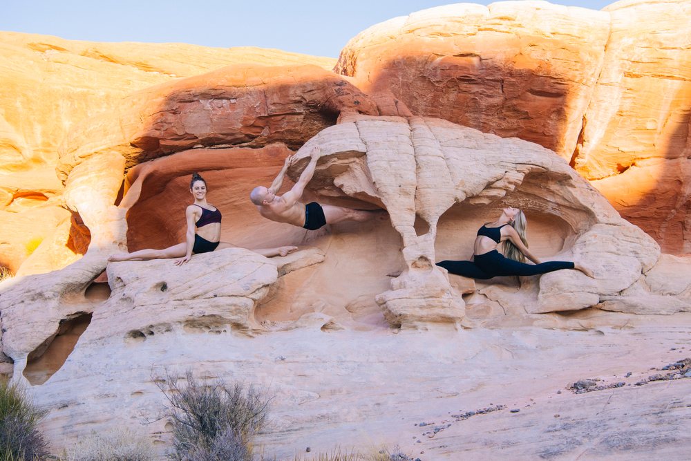 Three people practicing yoga and acrobatics on natural rock formations in a desert landscape, USA.