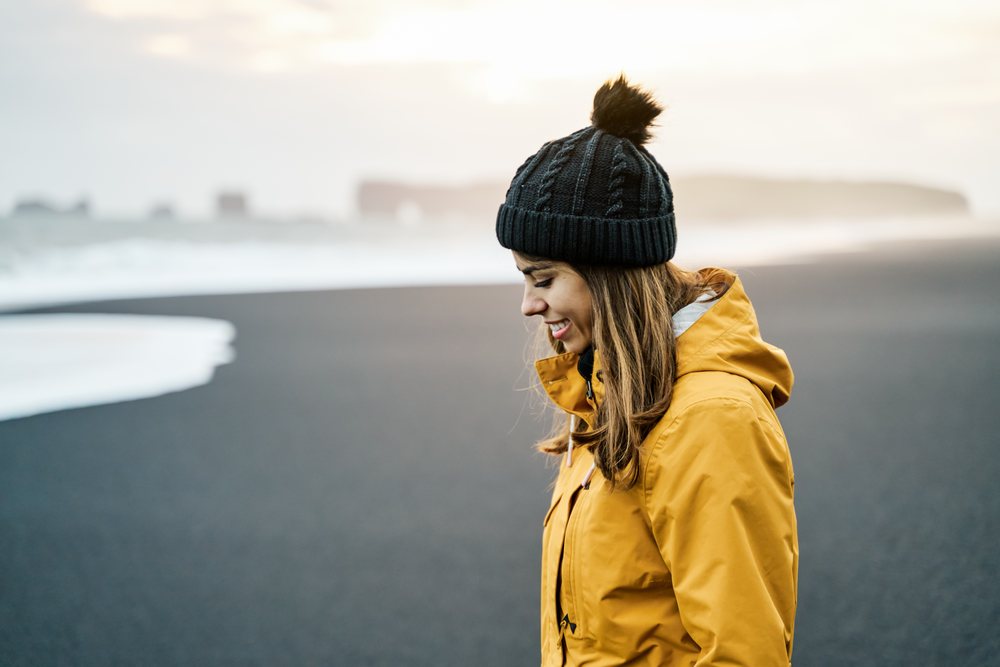 A white woman in a yellow jacket enjoys standing on a black sand beach
