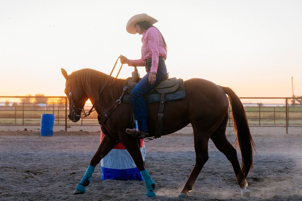 Woman riding a horse wearing blue leg boots with a western saddle in an outdoor arena at sunset, Southwest USA.