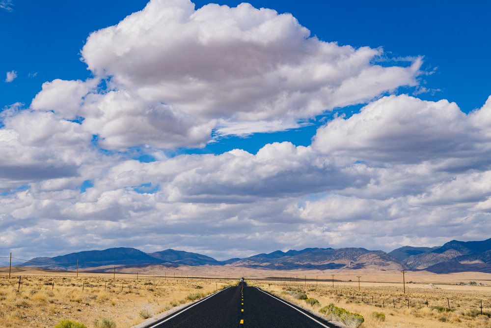 Open paved road with lane markings stretches through an arid desert landscape with mountains and scattered clouds, Nevada, USA.