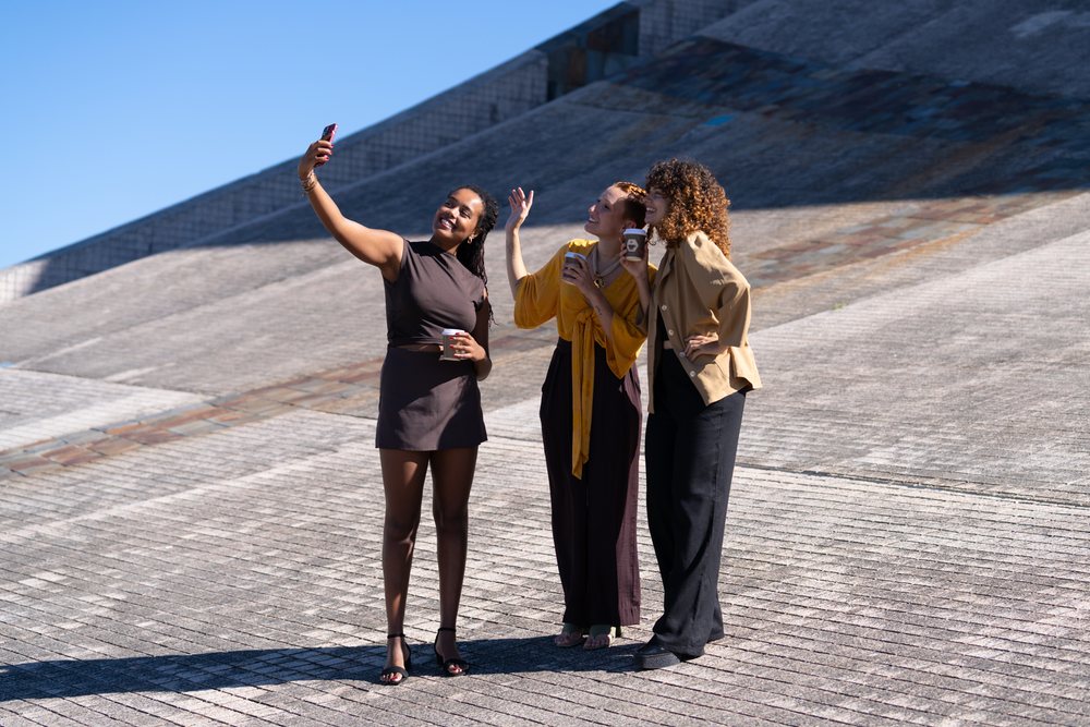 Three women outdoors in an urban area in Spain smiling and posing for a selfie while holding coffee cups on a sunny day.