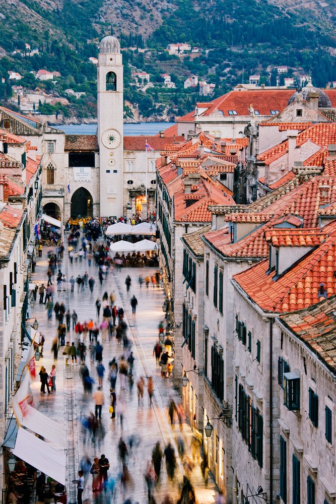 Crowded street in Dubrovnik's old city with stone buildings, red tile roofs, a clock tower, and outdoor dining by the Adriatic Sea.