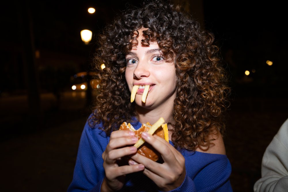 Woman with curly brown hair wearing a blue sweater playfully eats a cheeseburger and salted fries outdoors at night, Madrid, Spain.