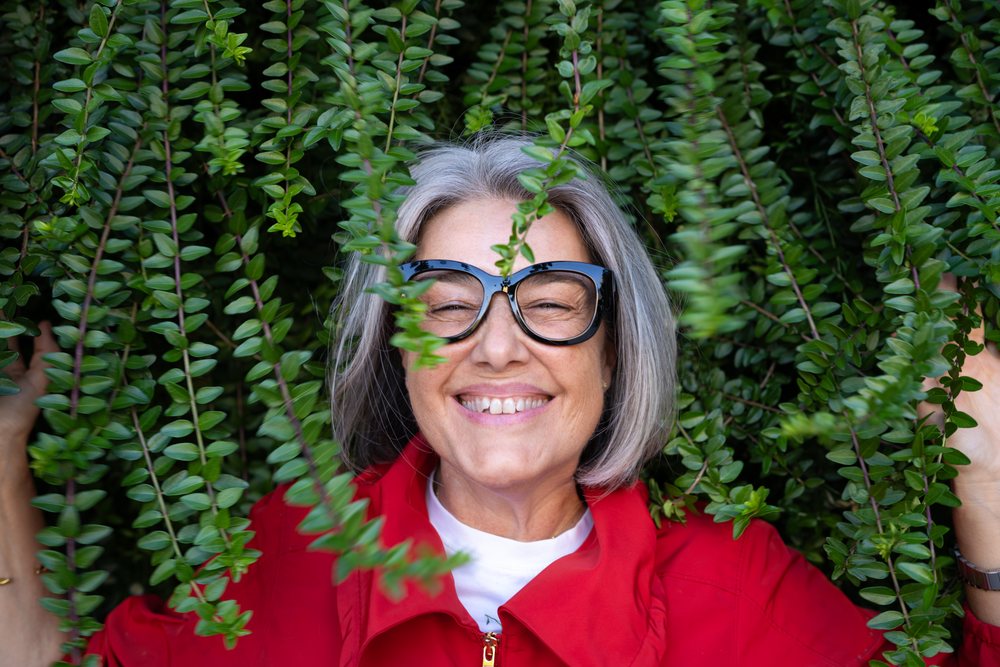 Smiling middle-aged woman with gray hair and black glasses wearing a red jacket among green leaves outdoors, Spain.