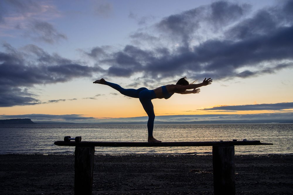 Person balancing in warrior III yoga pose on a wooden bench at sunset on the beach by the ocean, WA, USA.
