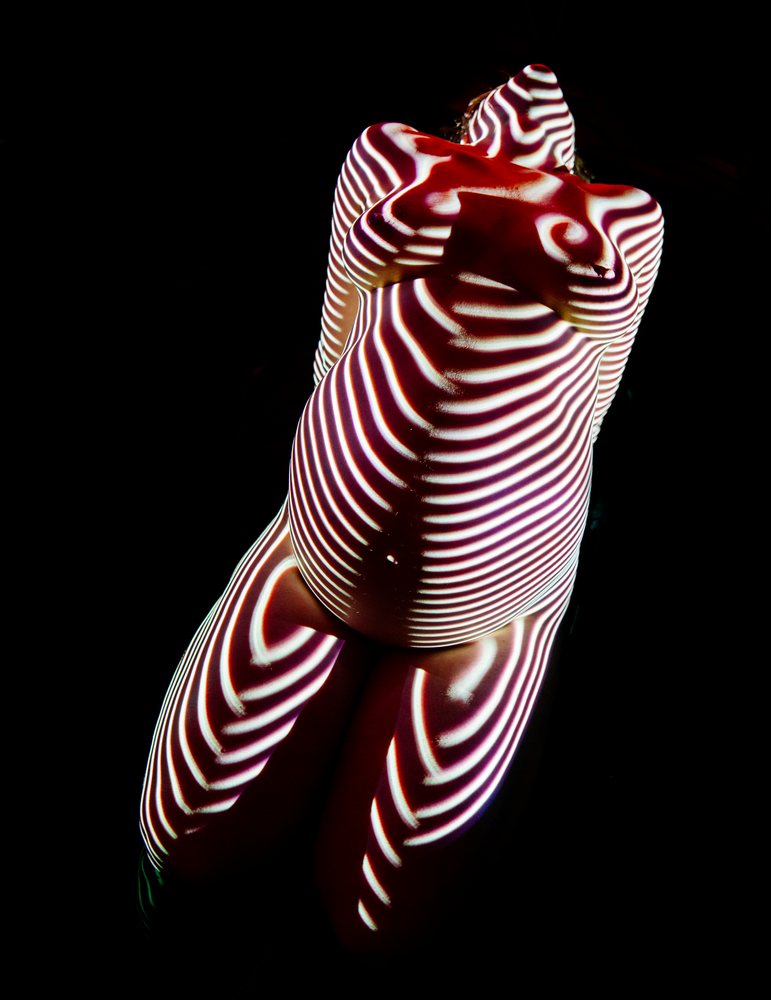Pregnant woman kneeling with striped light projected on her nude body against a dark background, pregnancy.