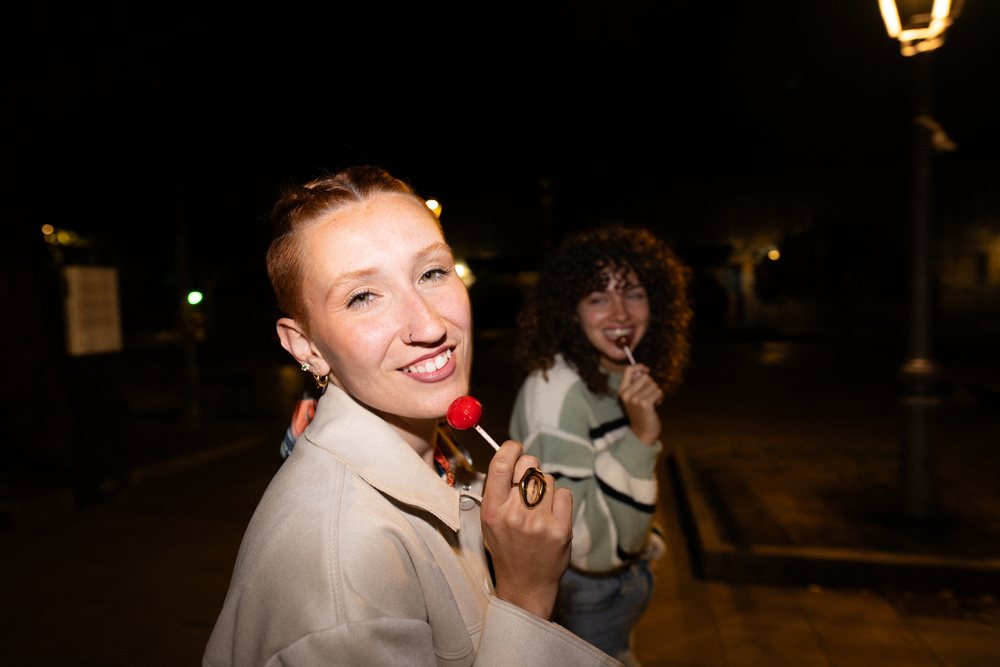 Two women holding lollipops and smiling outdoors at night in an urban street, friendship, Santiago De Compostela, Spain.