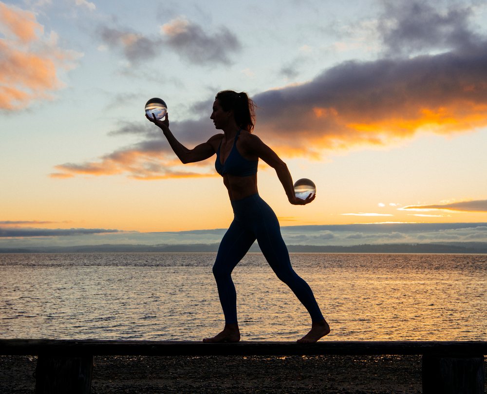 Woman barefoot in blue outfit holding glass spheres, balancing on a pier at sunset over the ocean, WA, USA.