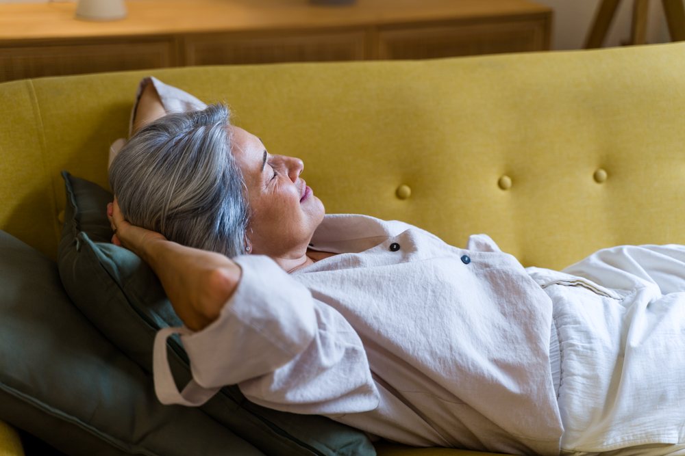Mature woman with gray hair rests with eyes closed on a yellow buttoned sofa indoors holding a green pillow, Spain.