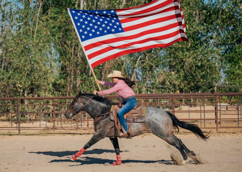 Female rider galloping a horse while holding an American flag in an outdoor sandy arena with trees and a metal fence, Southwest USA.