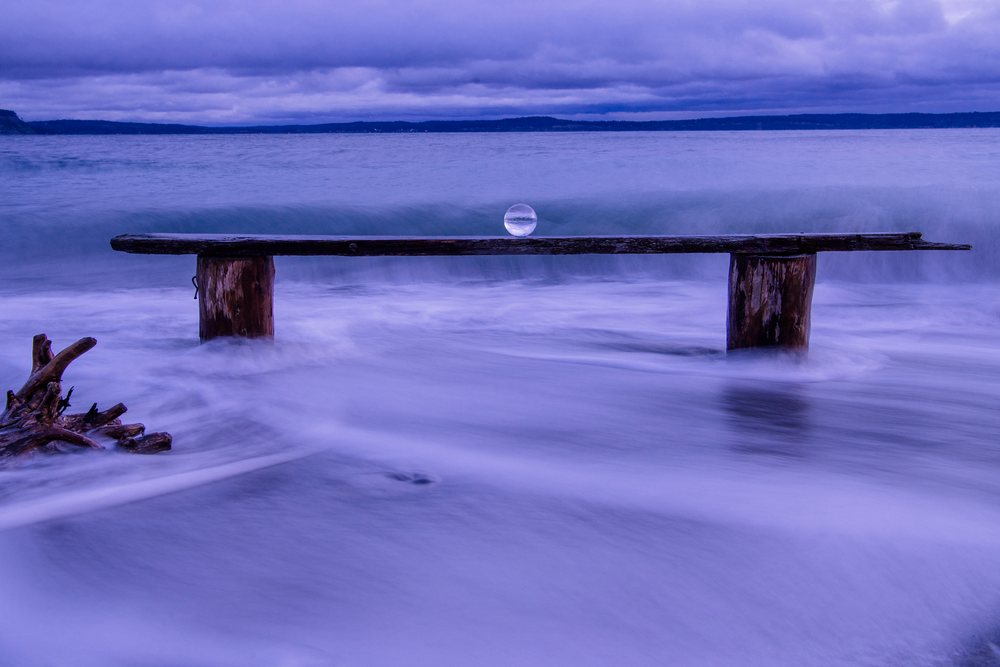 Wooden bench with glass sphere stands amid wave motion on the beach, Norwegian Point, WA, USA.