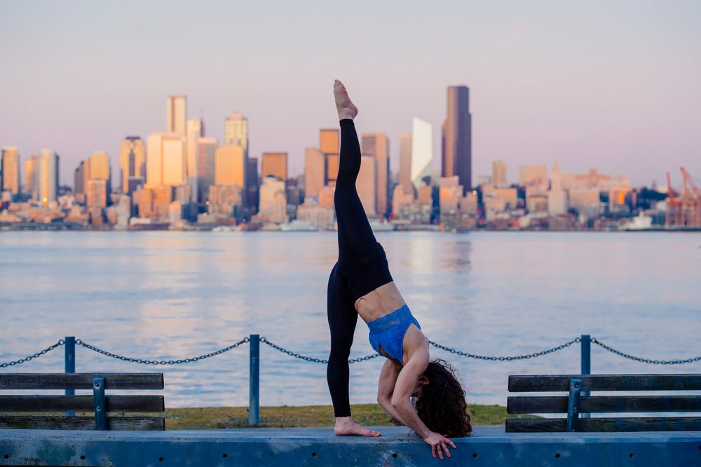 Woman barefoot is performing a yoga stretching pose on a waterfront promenade with a chain fence, wooden benches, and the Seattle skyline across calm ter at sunset, WA, USA.