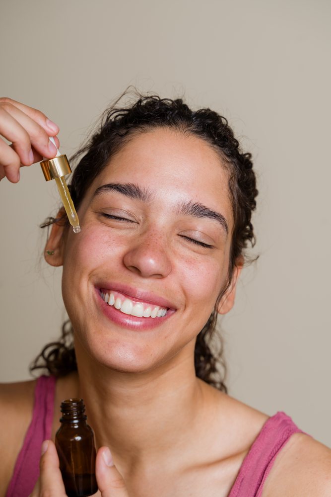 Woman with curly hair smiling and applying facial serum using a dropper from a brown bottle, beauty shoot, Spain.