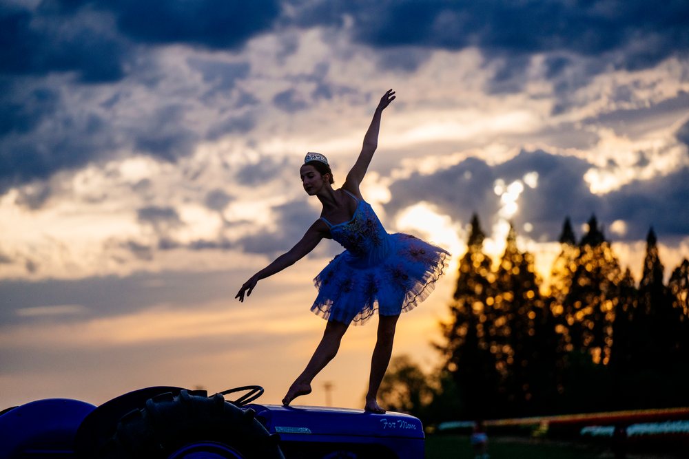 Ballet dancer wearing tutu and tiara performs a ballet pose barefoot while standing on a tractor outdoors at sunset, USA.