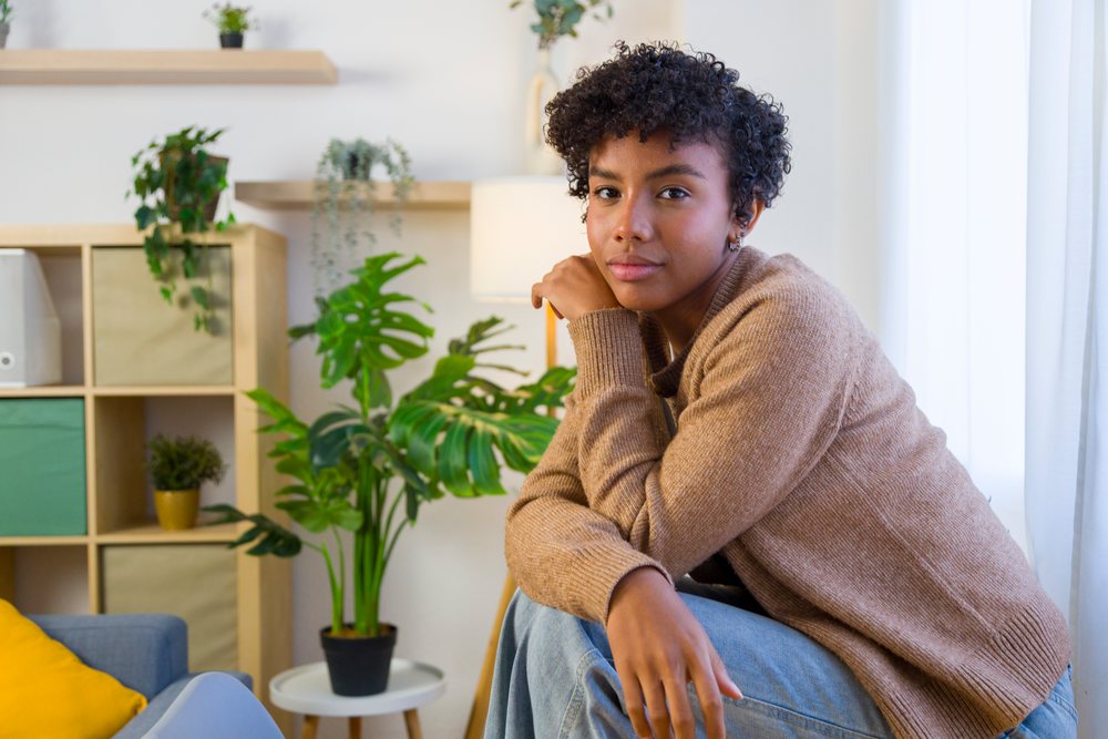 Young woman with curly hair wearing a brown sweater sitting indoors among plants and modern home decor.