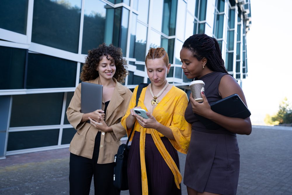 Three women stand outside a modern glass building, using smartphones, tablets, and a laptop, holding coffee and engaging in conversation, Spain.