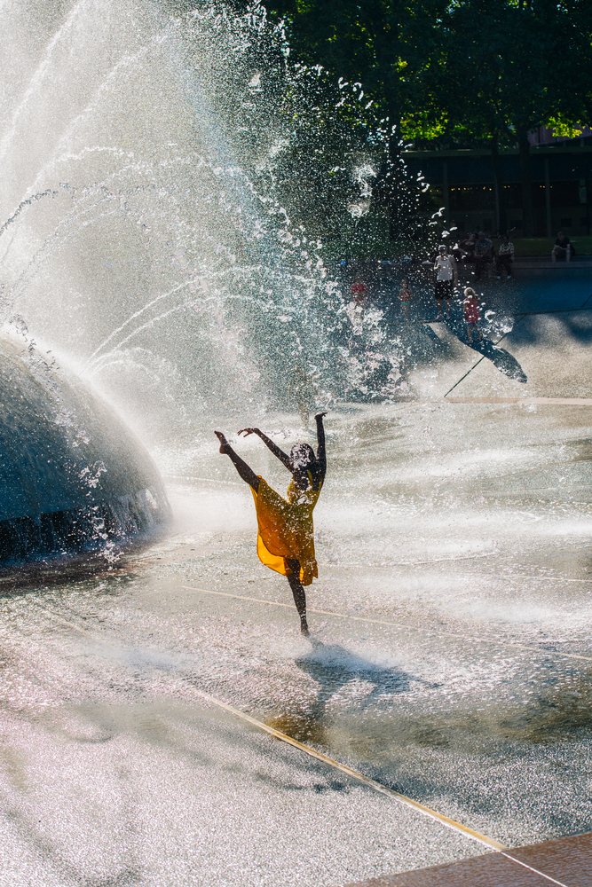 Dancer in a flowing yellow dress poses with one leg raised beneath large water jets of an outdoor fountain in a USA park.