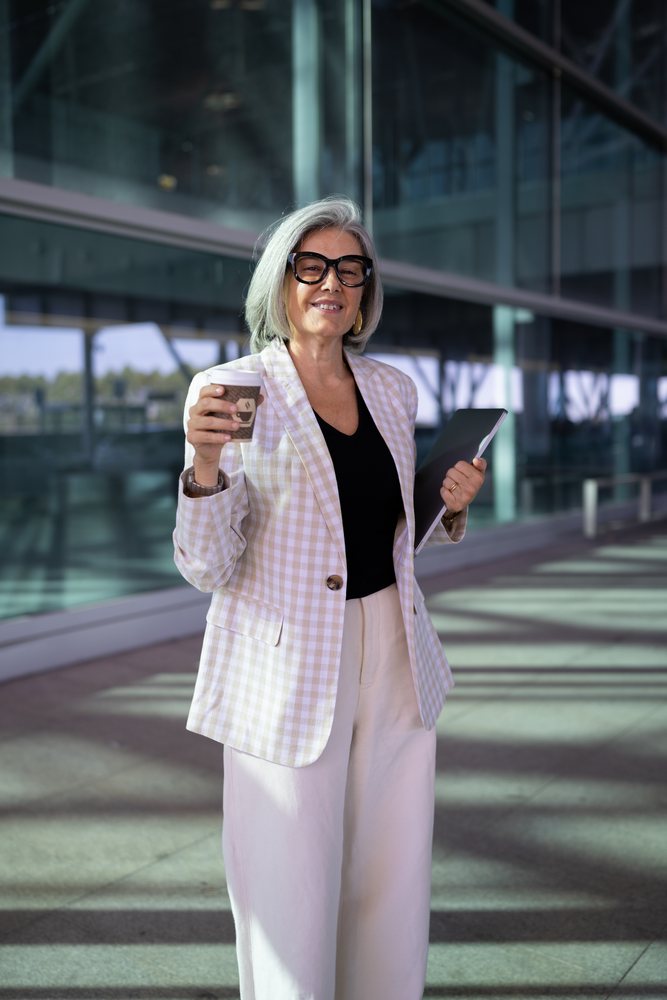 Businesswoman with gray hair and glasses holding a coffee cup and folder outside a modern glass building, Spain, Madrid.