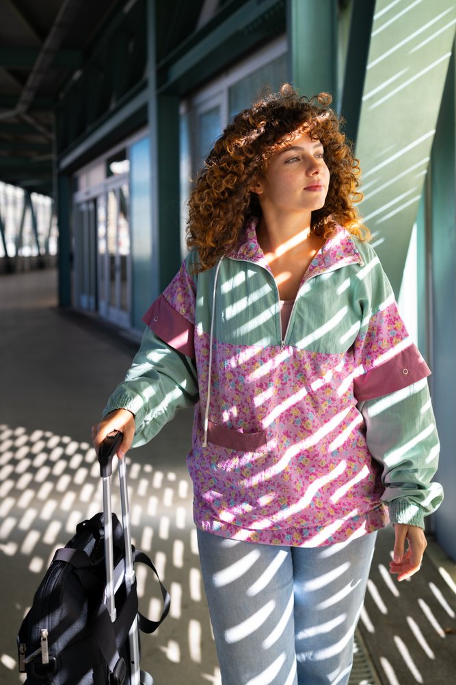 Woman with curly hair wearing pink jacket and jeans stands with suitcase in sunlit corridor, Spain.