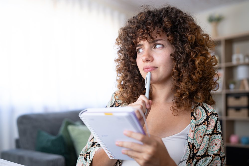 Girl holding a pen and notebook thoughtfully while sitting in a modern living room at home, Spain.