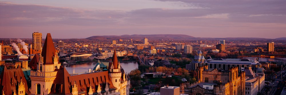 Cityscape at sunset showing a river flowing under a bridge with buildings and a park nearby.