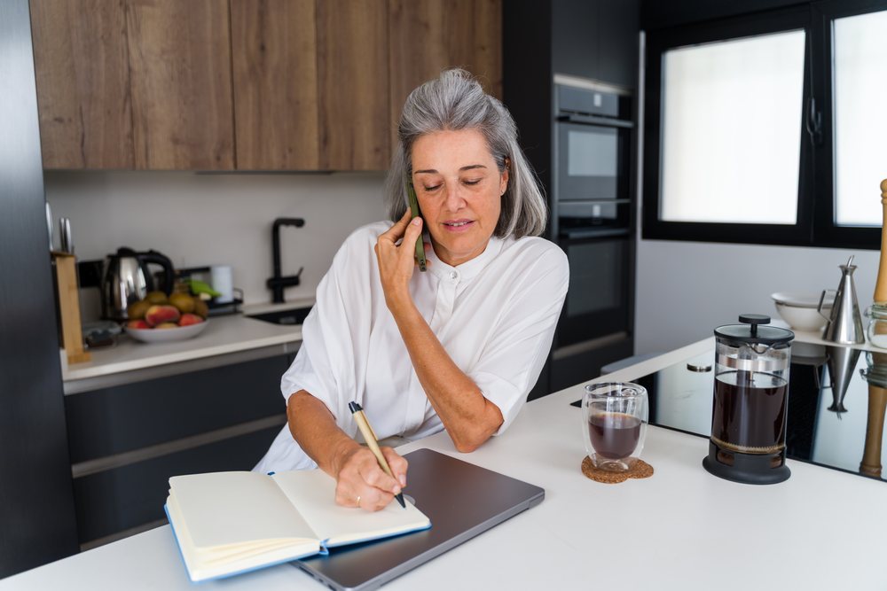 Mature woman writing in a notebook while talking on the phone in a modern kitchen, Spain.