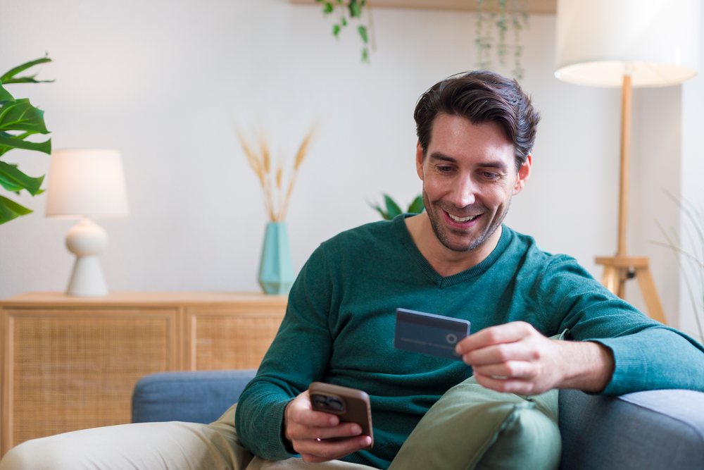 Smiling man holds a credit card and uses his smartphone while seated in a modern living room, technology, young adult, home.