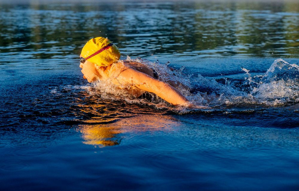 Swimmer wearing a yellow swim cap and goggles swimming freestyle while breathing in an open water lake, USA.