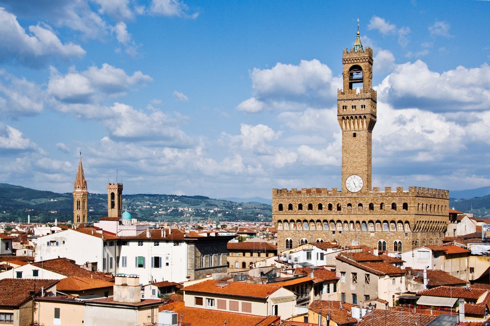 Palazzo Vecchio clock tower rises above Florence rooftops with Tuscan hills in the background under a blue sky, Italy.